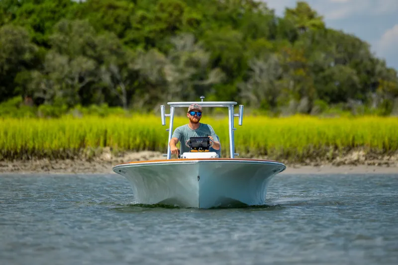 Slide: The Image of Man navigating 2023 Custom Carolina 18 Flats boat on calm water, grassy shoreline in background. - 2