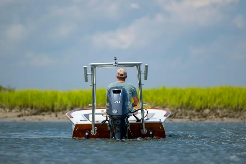 Slide: The Image of 2023 Custom Carolina 18 Flats boat on water with person steering, grassy shoreline in background. - 6