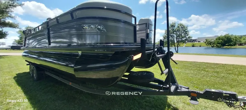 Slide: The Image of 2023 Regency 250 LE3 Sport pontoon boat on trailer, parked on grass by a lake. - 21