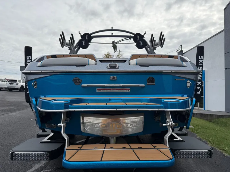 Slide: The Image of 2020 Malibu Wakesetter 23 MXZ boat, rear view, blue with teak accents, parked outdoors. - 8
