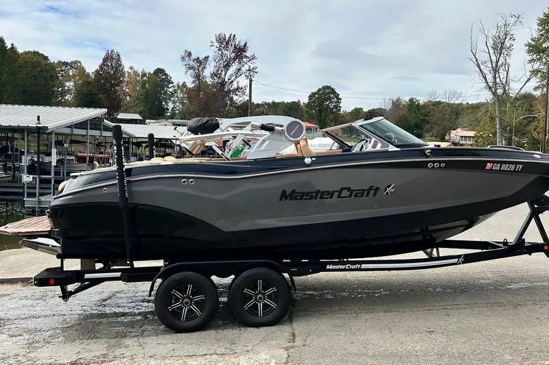 Slide: The Image of 2020 MasterCraft X22 boat on trailer, parked near a marina with trees in the background. - 2