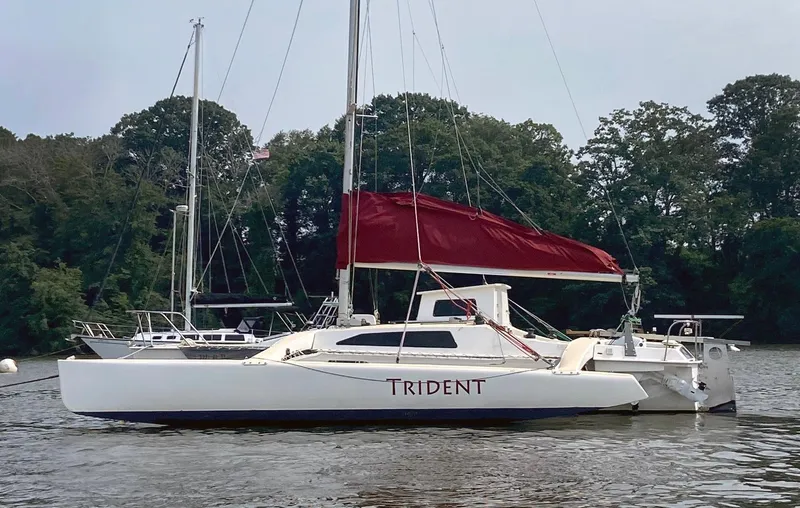 Slide: The Image of Corsair F27 sailboat from 1990 with red sail, named "Trident," on a calm river. - 2