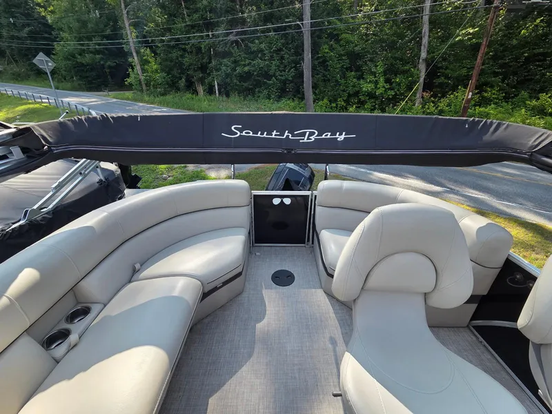 Slide: The Image of 2018 South Bay 523 pontoon boat interior with beige seating and canopy, surrounded by lush greenery. - 17