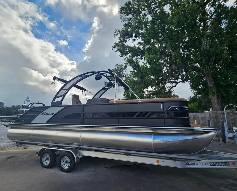 Slide: The Image of 2026 Bentley Pontoons Elite 253 Swingback on trailer, parked near trees and cloudy sky. - 4