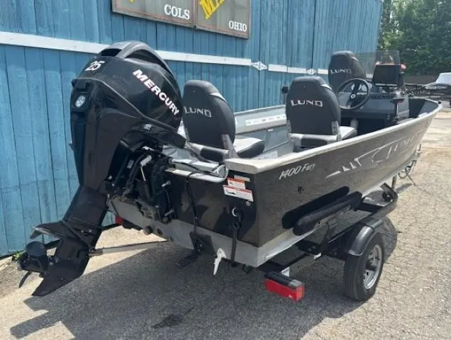 Slide: The Image of 2013 Lund 1400 Fury SS boat with Mercury engine, parked on a trailer near a blue building. - 2