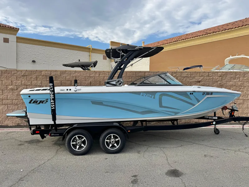 Slide: The Image of 2019 Tigé R21 boat on trailer, light blue and white design, parked outdoors. - 0