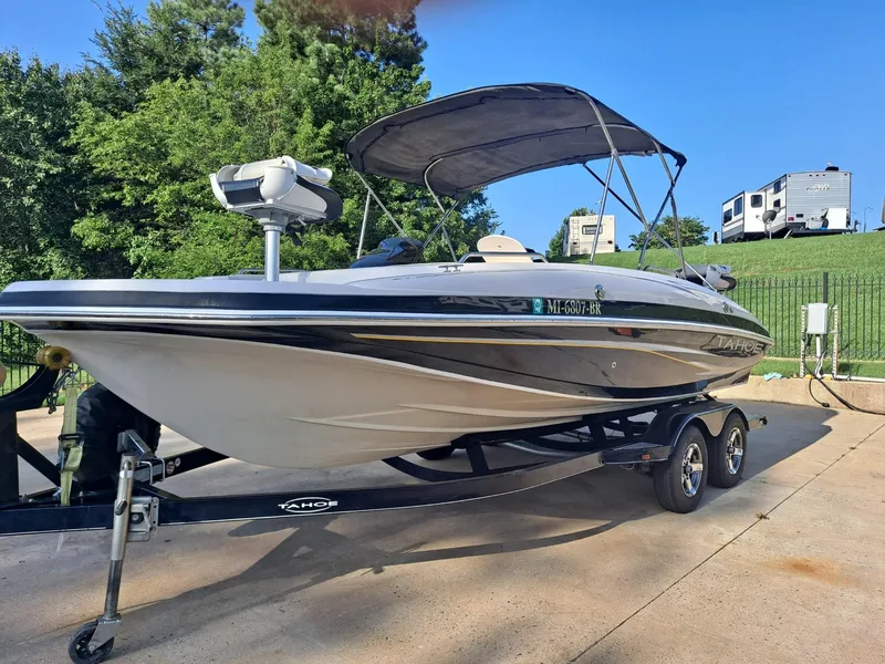 Slide: The Image of 2010 Tahoe 195 Deckboat on trailer, parked outdoors with trees and RVs in the background. - 1