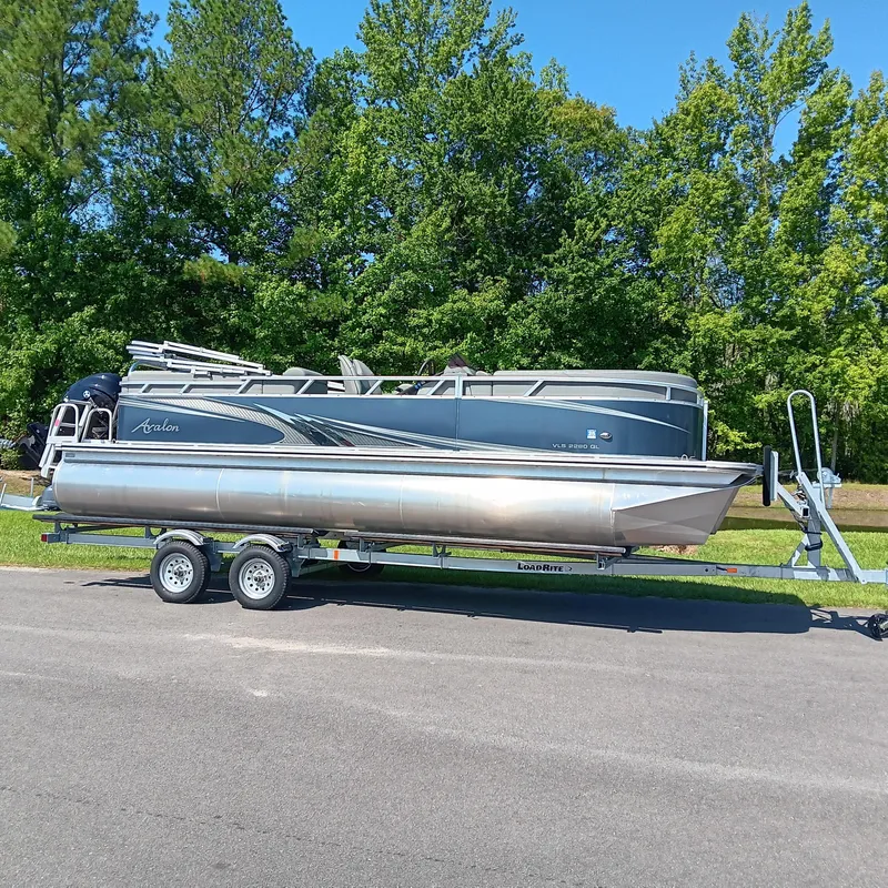 Slide: The Image of 2022 Avalon VLS Quad Lounge pontoon boat on trailer, parked on road with trees in background. - 1