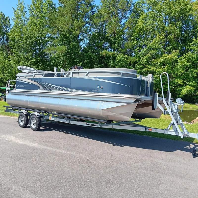 Slide: The Image of 2022 Avalon VLS Quad Lounge pontoon boat on trailer, surrounded by lush greenery. - 2