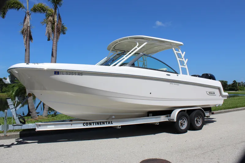 Slide: The Image of 2018 Boston Whaler 270 Vantage boat on trailer, parked near palm trees under clear blue sky. - 2
