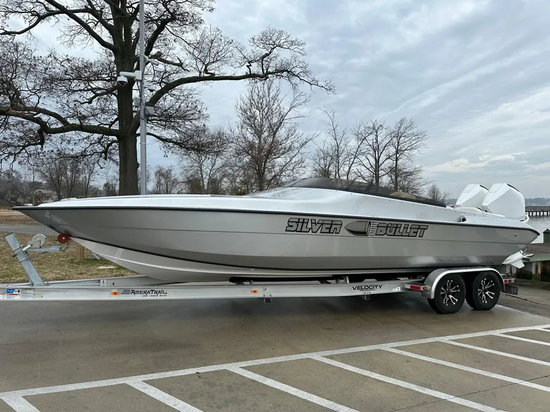 Slide: The Image of 2022 Velocity 290 SC "Silver Bullet" speedboat on trailer, parked near trees and cloudy sky. - 14