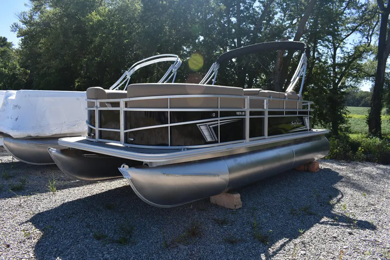 Slide: The Image of 2024 SunChaser Vista 20 Fish pontoon boat on gravel lot with trees in background. - 3