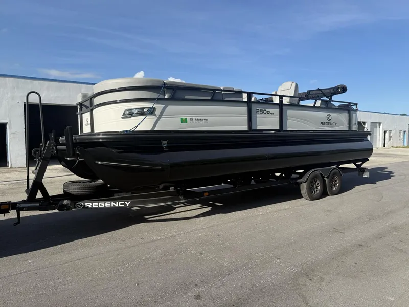 The Image of 2024 Regency 250 DL3 pontoon boat on trailer, parked outdoors under clear sky. - 0