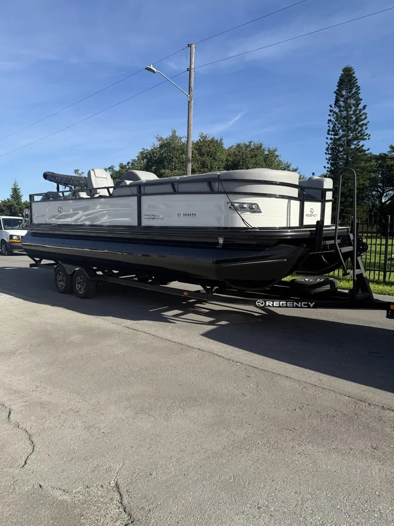 Slide: The Image of 2024 Regency 250 DL3 pontoon boat on trailer, parked outdoors under clear sky. - 2