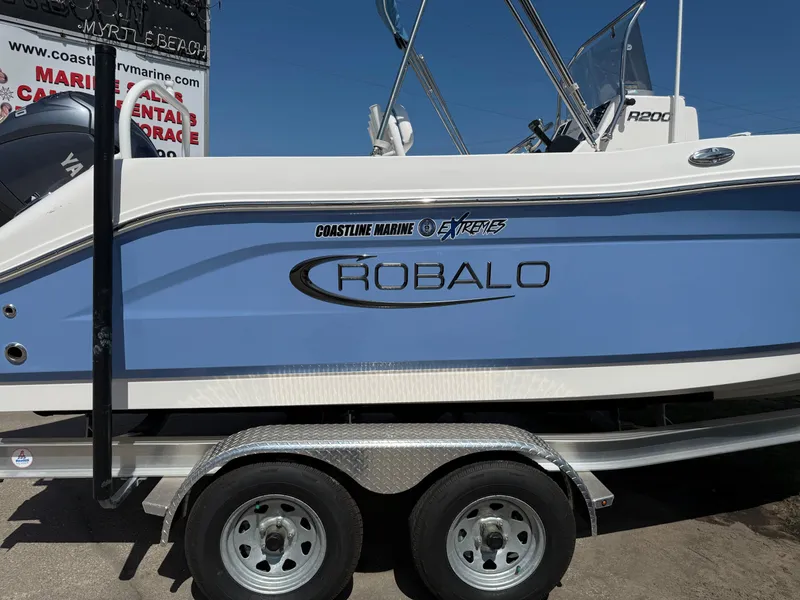 The Image of 2025 Robalo R200 Center Console boat on trailer, blue and white design. - 0
