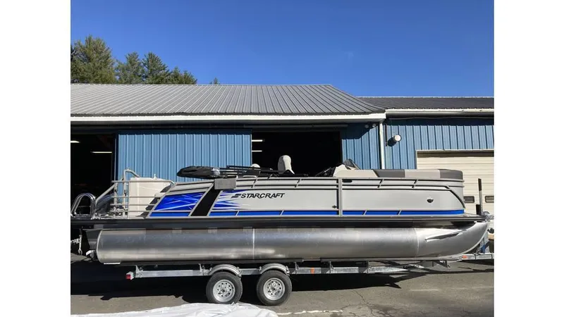 Slide: The Image of 2025 Starcraft CX 23 RL pontoon boat on trailer, parked outside blue building. - 6