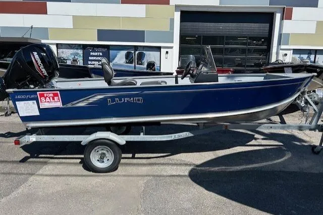 Slide: The Image of 2026 Lund 1600 Fury SS boat for sale, blue exterior, on trailer, outside dealership. - 2