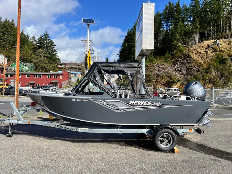 The Image of 2025 Hewescraft 180 Sportsman boat on trailer, parked outdoors with trees and buildings in background. - 0