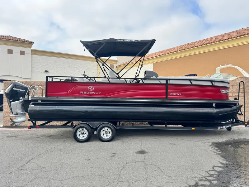 The Image of 2023 Regency 250 DL3 pontoon boat on trailer, red and black design, parked outdoors. - 0