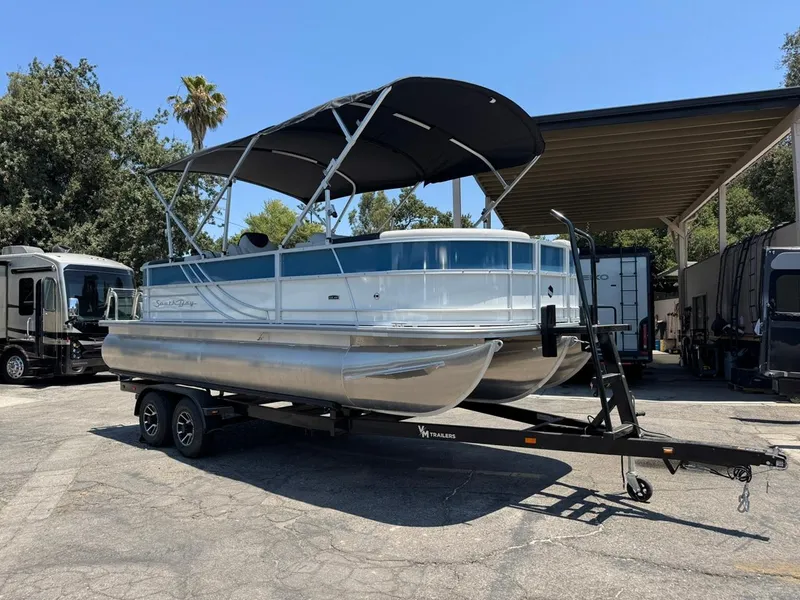 Slide: The Image of 2025 South Bay 222 LE pontoon boat on trailer, parked outdoors under clear sky. - 2