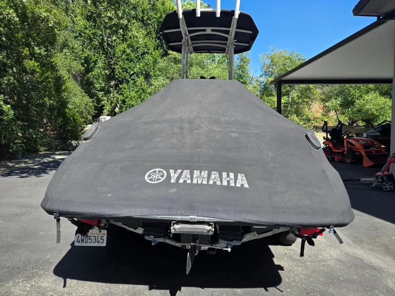 Slide: The Image of 2022 Yamaha Boats 210 FSH Deluxe covered, parked outdoors with trees and equipment nearby. - 5