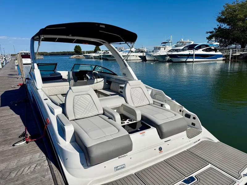 Slide: The Image of The image shows a 2026 Sea Ray SDX 290 boat docked. It features sleek gray and white upholstery with a black sunshade. Other boats and calm water are visible in the background. - 2