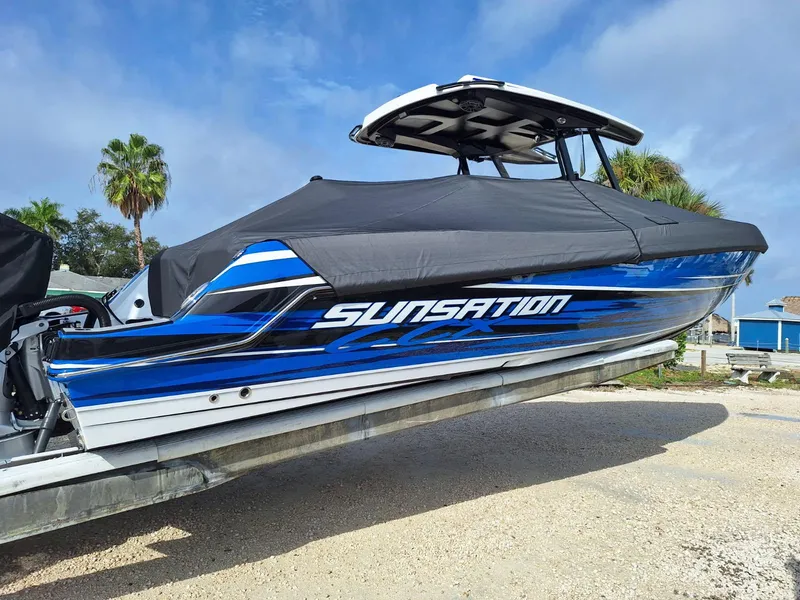 Slide: The Image of 2023 Sunsation 32 CCX boat with cover, displayed on a trailer, palm trees in background. - 16