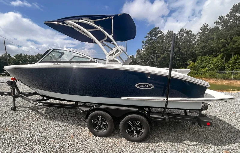 Slide: The Image of 2018 Cobalt CS23 boat on trailer, blue and white, parked on gravel with trees in background. - 7