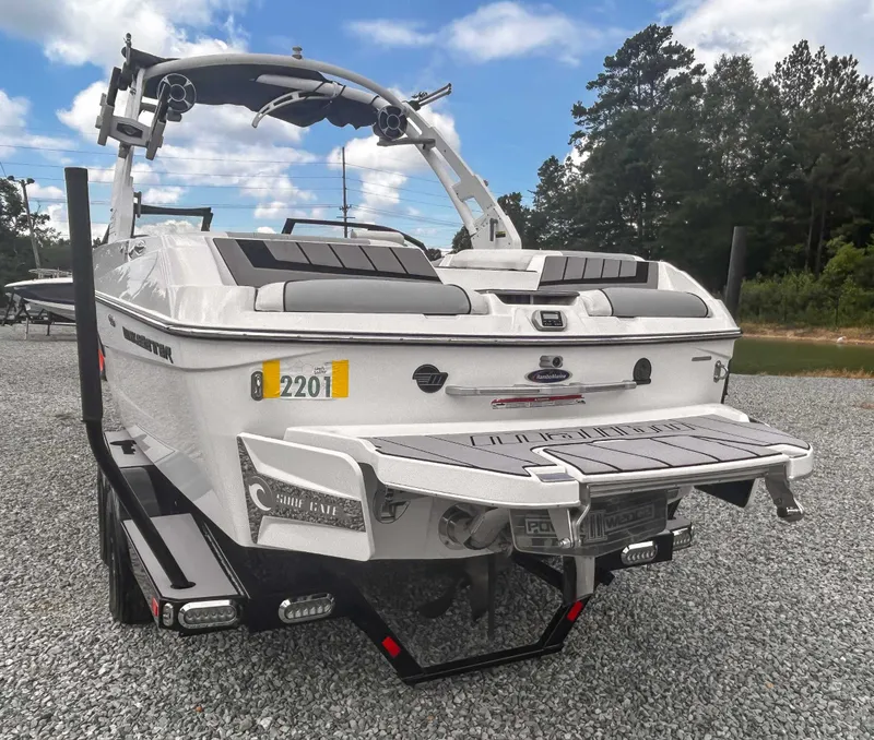 Slide: The Image of 2020 Malibu 23 MXZ boat on trailer, parked on gravel, with trees in background. - 7