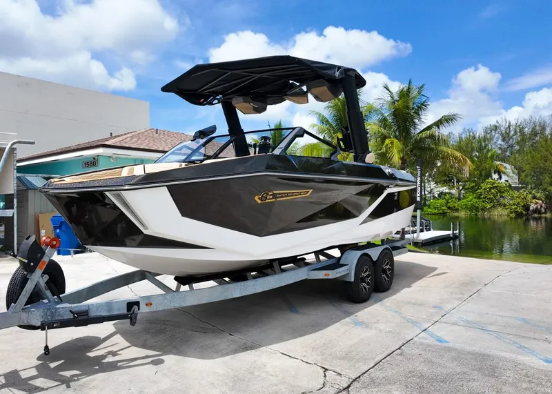 Slide: The Image of 2025 Nautique G23 boat on trailer, parked near water, under clear blue sky. - 2