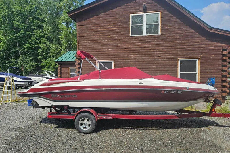 The Image of 2008 Bryant 233 boat with red cover on trailer, parked by wooden cabin. - 0