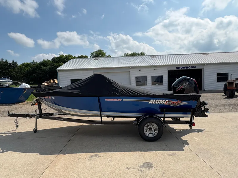 Slide: The Image of 2018 Alumacraft Voyageur 175 Sport boat with cover, parked outside a showroom. - 31