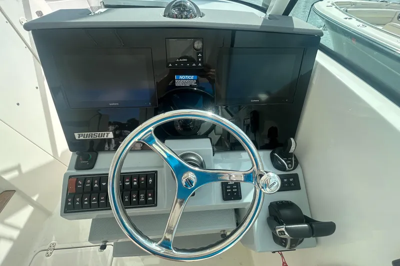 Slide: The Image of 2023 Pursuit DC326 boat helm with steering wheel, controls, and marina view. - 13