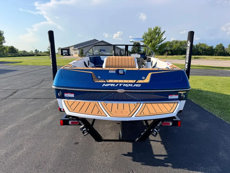 Slide: The Image of 2025 Nautique Ski 200 boat, blue exterior, parked on pavement with green grass and clear sky. - 4