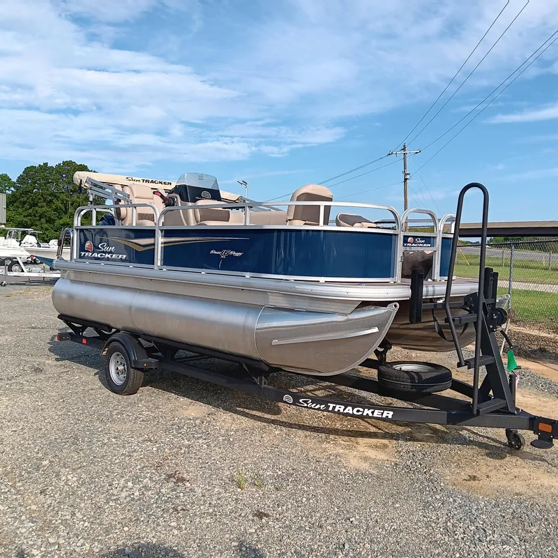 Slide: The Image of 2025 Sun Tracker Bass Buggy 16 XL Select pontoon boat on trailer, parked outdoors. - 2
