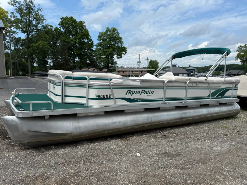 Slide: The Image of 1999 AquaPatio 270fe pontoon boat parked on gravel, featuring green and white design. - 1