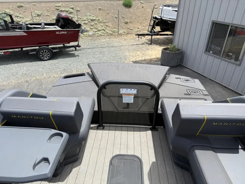Slide: The Image of 2025 Manitou Explore boat interior with seating and table, parked near a metal building. - 12