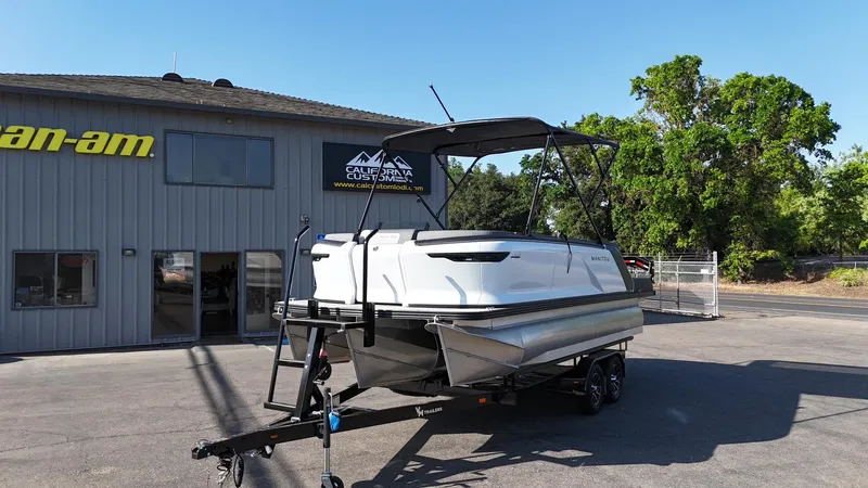 The Image of 2025 Manitou Cruise boat on trailer outside dealership, clear sky, trees in background. - 1