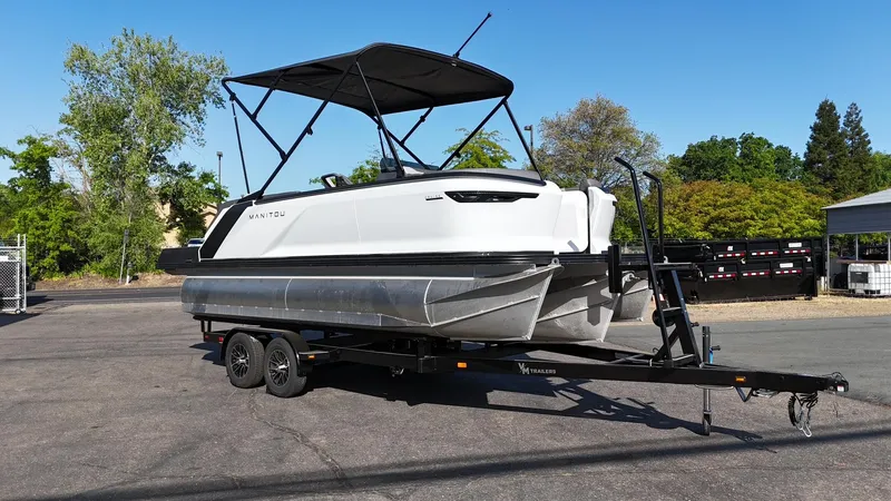 Slide: The Image of 2025 Manitou Cruise pontoon boat on trailer, parked outdoors under clear blue sky. - 2