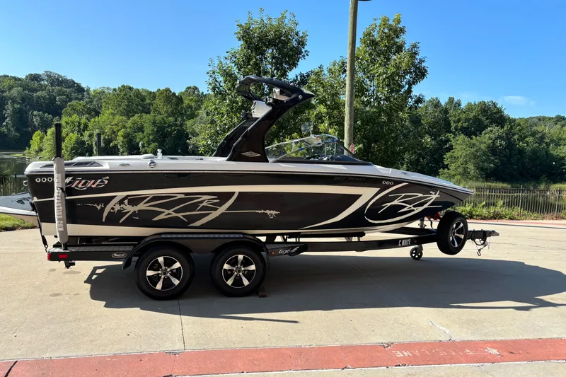 The Image of 2014 Tigé RZR boat on trailer, parked near a scenic lake with lush greenery. - 0