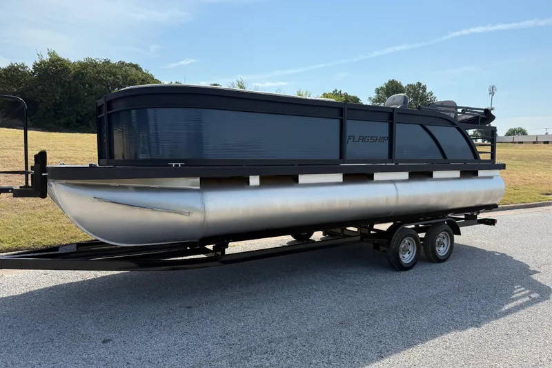 Slide: The Image of 2025 Flagship E22RF Sport Toon pontoon boat on trailer, parked outdoors. - 0