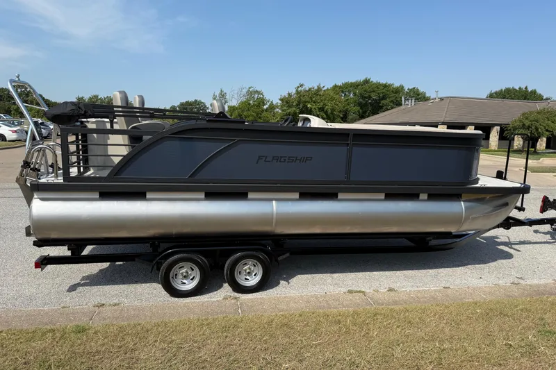 Slide: The Image of 2025 Flagship E22RF Sport Toon pontoon boat on trailer, parked outdoors. - 18
