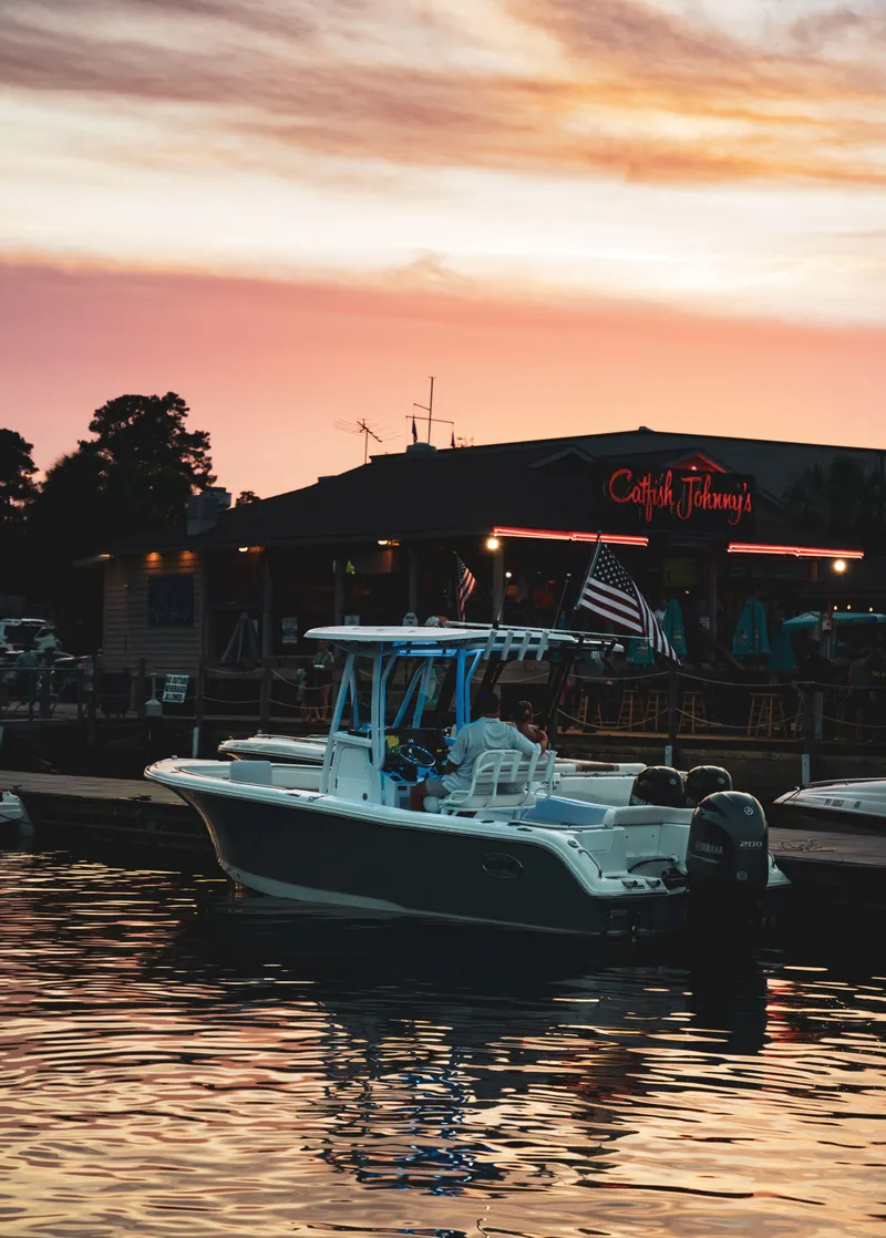 Slide: The Image of 2024 Sea Hunt Ultra 234 boat docked at sunset near a waterfront restaurant. - 30