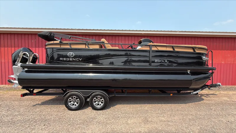The Image of 2025 Regency 230 LE3 pontoon boat on trailer, parked by red building. - 0