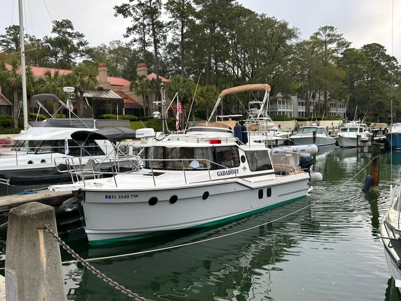 Slide: The Image of 2024 Ranger Tugs R-29 CB docked in a marina, surrounded by trees and other boats. - 2