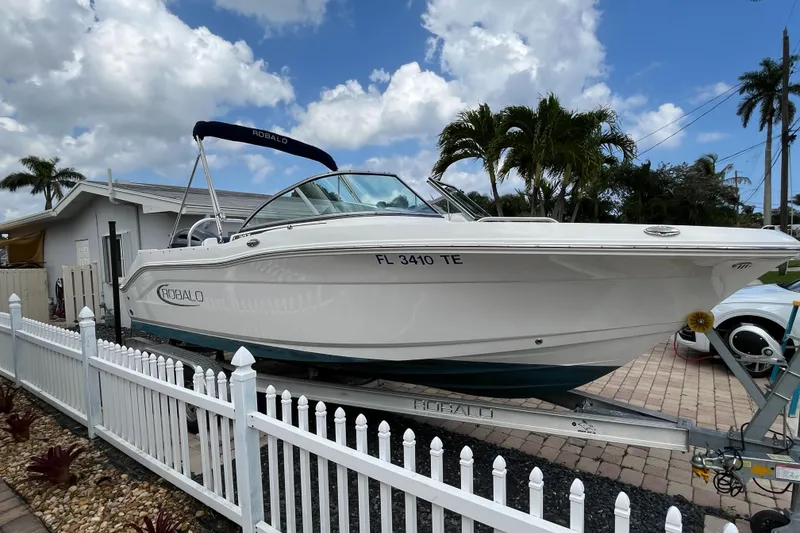 Slide: The Image of 2021 Robalo 207R boat on trailer, parked beside a white fence under a cloudy sky. - 1