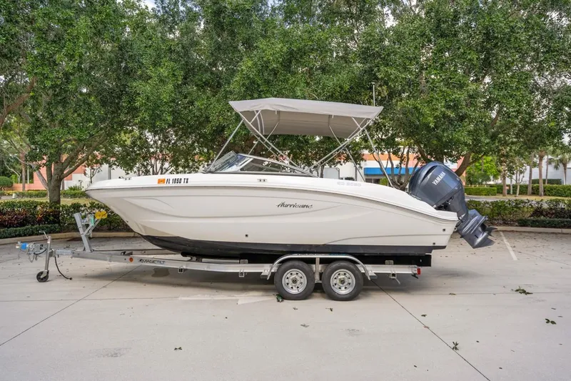 Slide: The Image of 2025 Hurricane SunDeck 217 OB boat on trailer, parked outdoors with trees in background. - 0