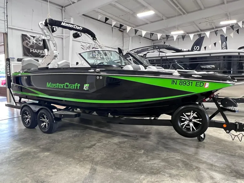 The Image of 2015 MasterCraft X25 boat with green accents on a trailer in a showroom. - 0