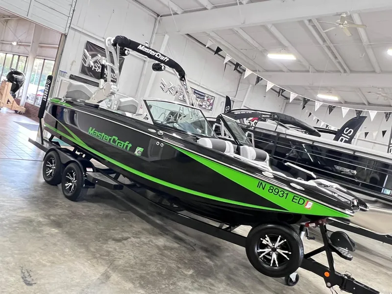 Slide: The Image of 2015 MasterCraft X25 boat with green accents on a trailer in a showroom. - 1