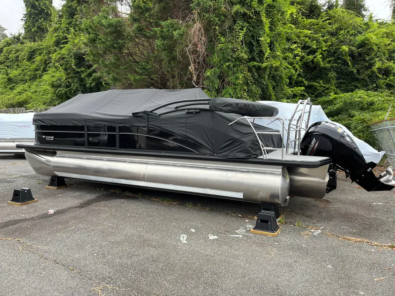 Slide: The Image of 2026 Harris Cruiser 230 pontoon boat covered, parked on pavement with lush greenery background. - 2